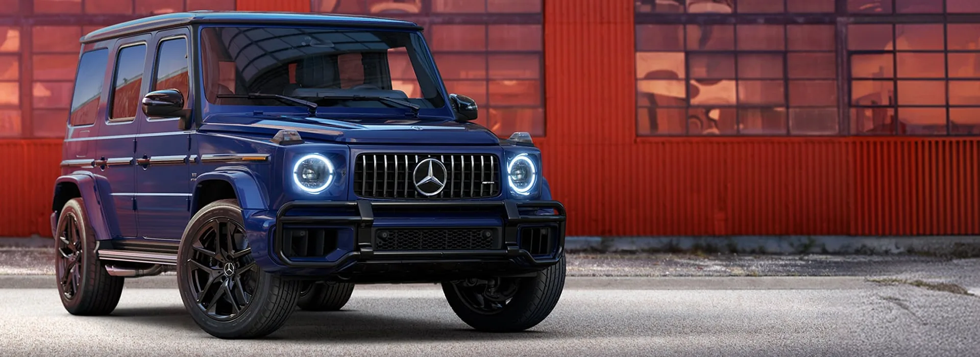2027 Mercedes-AMG G 63 in blue, shown from the front three-quarter angle against a red industrial backdrop
