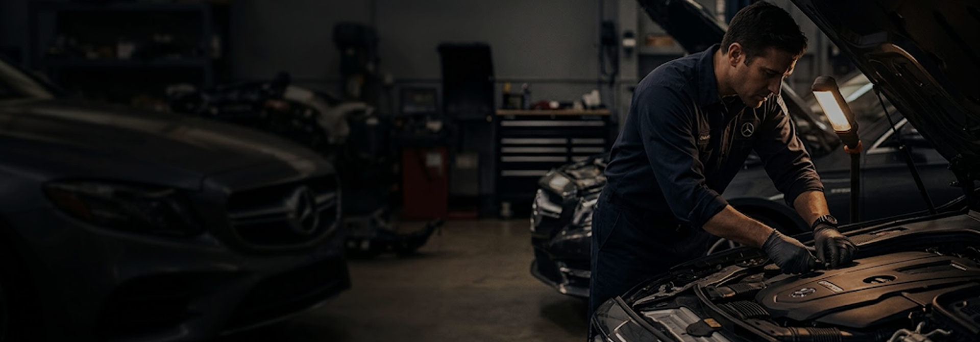 Mercedes-Benz technician in black gloves working under the open hood of a Mercedes-Benz in a dimly lit service bay