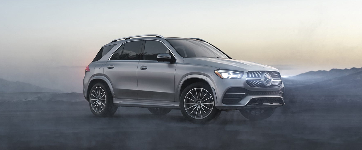 Silver Mercedes-Benz GLE mid-size luxury SUV in a front three-quarter view, parked on a ridge at dusk with mist and mountains behind it