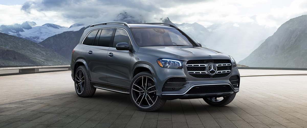 Grey Mercedes-Benz GLS full-size luxury SUV in a front three-quarter view on a paved overlook, with snow-capped mountains and breaking cloud behind it